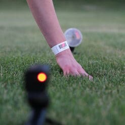 A person wearing a Dashr Barcode Wrist Band reaches down to touch grass, while small devices emitting red lights are visible, likely for measurement or monitoring purposes.
