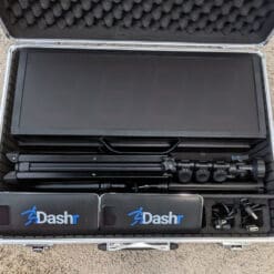 A Dashr Display Pack rests in an open carrying case on a carpeted floor, showcasing its components: a large digital display, tripods, sensor devices, and accessories neatly arranged inside.