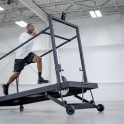 In an industrial-style gym, a person runs on the SHREDmill, a large inclined treadmill, beneath high ceilings and bright lighting. They sport a white shirt, black shorts, and black athletic shoes.