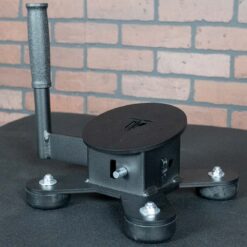 The Spring Ankle, a black three-legged device with a vertical handle, round base, leg bolts, and a slot on its main body, sits on a dark surface in front of a brick wall.