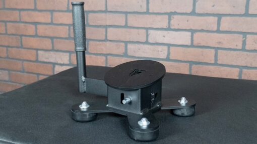 The Spring Ankle, a black three-legged device with a vertical handle, round base, leg bolts, and a slot on its main body, sits on a dark surface in front of a brick wall.
